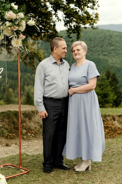 Um casal de idosos fica perto de um elegante arco de casamento debaixo de uma árvore contra o pano de fundo das montanhas, velho noivo e noiva