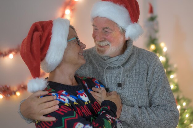 Um casal de idosos sorridente usando chapéus de Papai Noel em casa