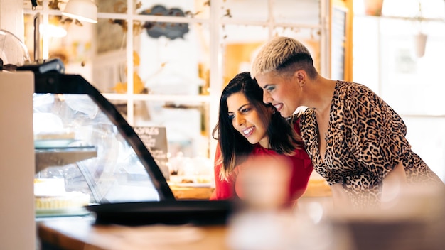 Foto um casal de lésbicas olha para uma vitrine e escolhe quais bolos levar em um café