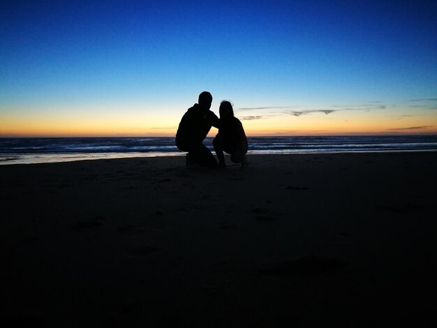 Um casal de silhuetas agachado na praia contra o céu azul ao anoitecer