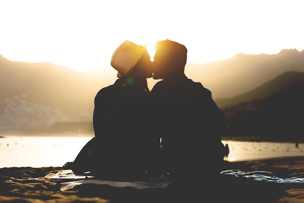 Um casal de siluetas a beijar-se enquanto estão sentados ao lado do lago durante o pôr-do-sol