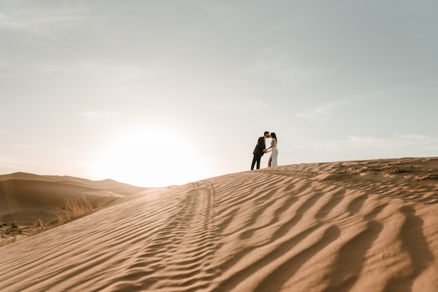 Um casal em suas roupas de casamento em cima de uma duna do deserto ao amanhecer