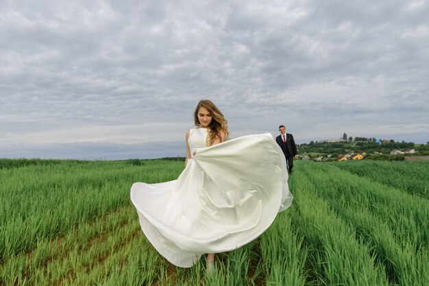 Um casal em um traje de casamento fica em um campo verde em uma vila ao pôr do sol, a noiva e o noivo. A noiva está girando em seu vestido. O noivo a admira.