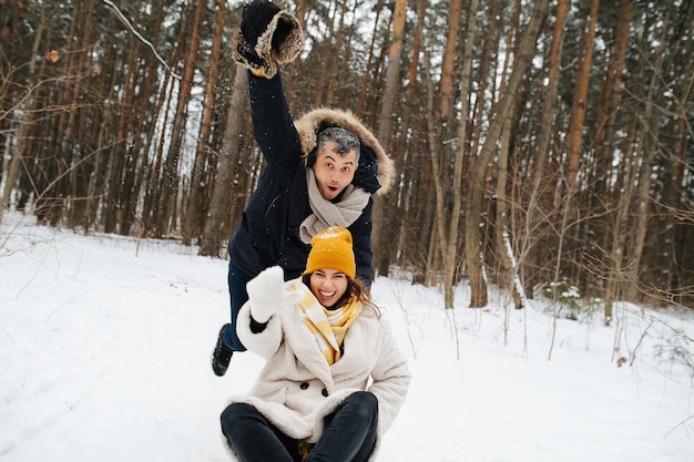 Um casal em uma floresta de inverno se diverte em um dia de folga, o marido empurra o trenó com a esposa. Ele tirou o boné e mostra o caminho