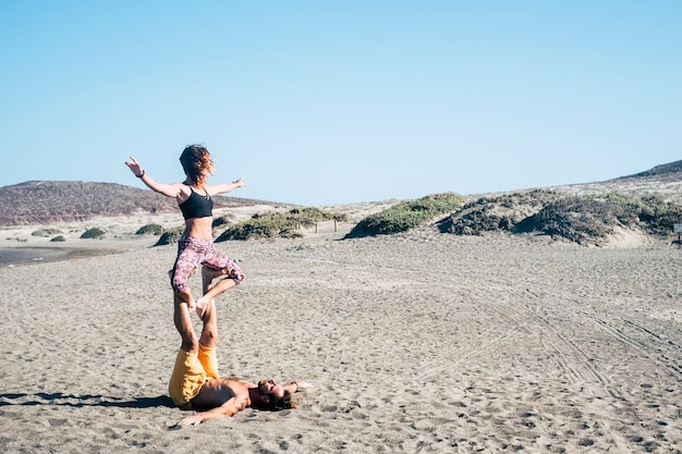 Um casal fazendo acroyoga ou ioga na praia em silêncio para se concentrar e não cair - na areia e com a areia e as pedras ao fundo