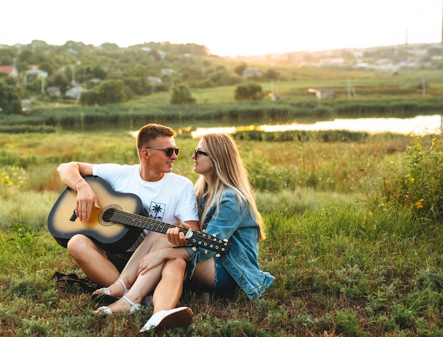 Um casal feliz, apaixonado por óculos de sol, tocando violão e regozijando-se ao pôr do sol.