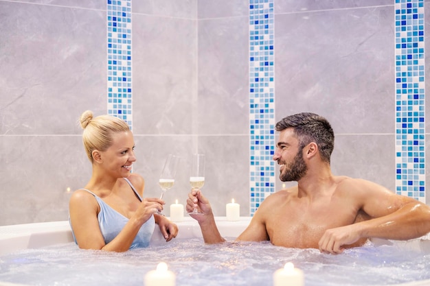 Um casal feliz está sentado na banheira de hidromassagem no centro de spa e brindando com champanhe