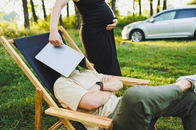 Um casal feliz relaxa em cadeiras dobráveis perto do carro um homem usa um laptop para o trabalho ou verificar redes sociais sua esposa trata-o de um croissant mulher grávida com homem na natureza