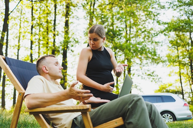 Um casal feliz relaxa em cadeiras dobráveis perto do carro um homem usa um laptop para o trabalho ou verificar redes sociais sua esposa trata-o de um croissant mulher grávida com homem na natureza