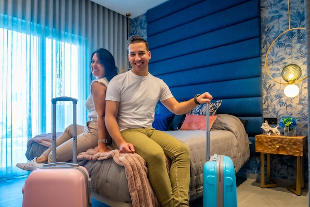 Foto um casal feliz sentado na cama de um fato de hotel cercado por bagagens desembaladas