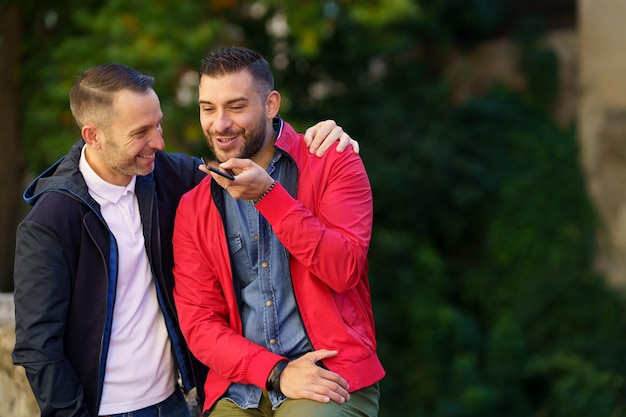 Foto um casal gay sorridente a falar ao telemóvel ao ar livre.