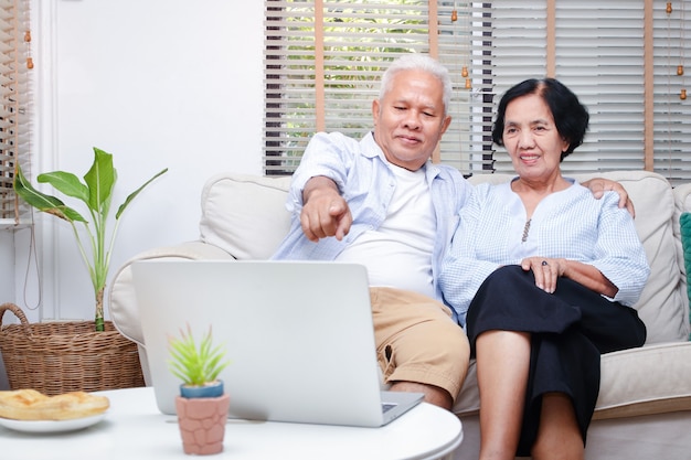 Um casal idoso de asiáticos assiste mídia online em seu laptop na sala de estar de casa.