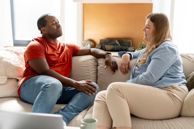 Um casal multiétnico feliz sentado no sofá aconchegante da sala de estar e olhando um para o outro.