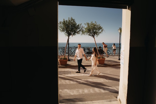 Um casal passeando no calçadão em frente a uma janela com o mar ao fundo.