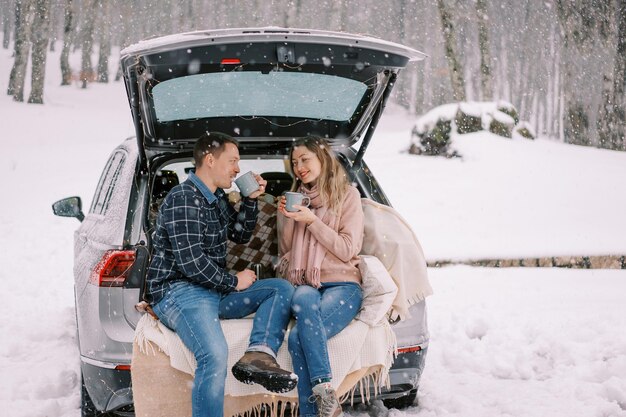 Um casal sorridente bebe café de copos enquanto está sentado no porta-malas aberto de um carro sob nevasca no