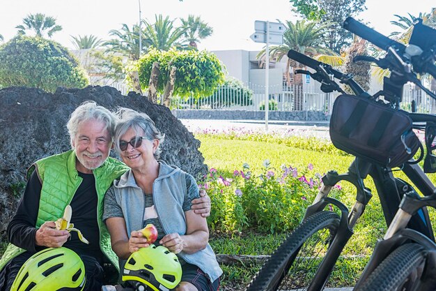 Um casal sorridente de ciclistas faz uma pausa no parque comendo algumas frutas sentadas no chão