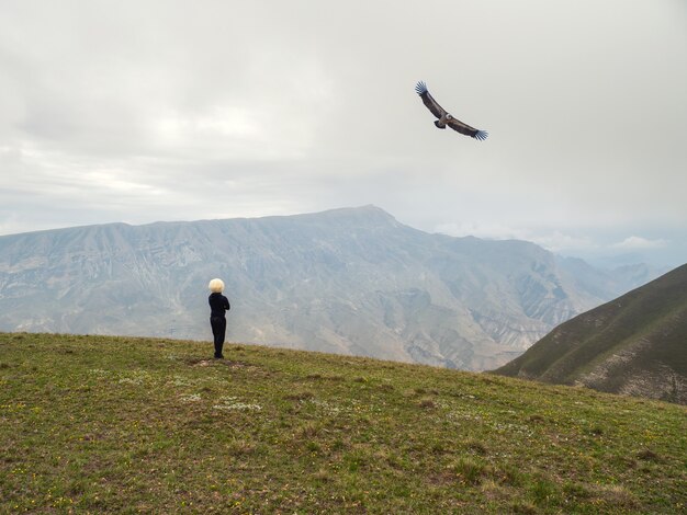 Um cavaleiro com um boné branco e uma águia voadora no fundo de altas montanhas.