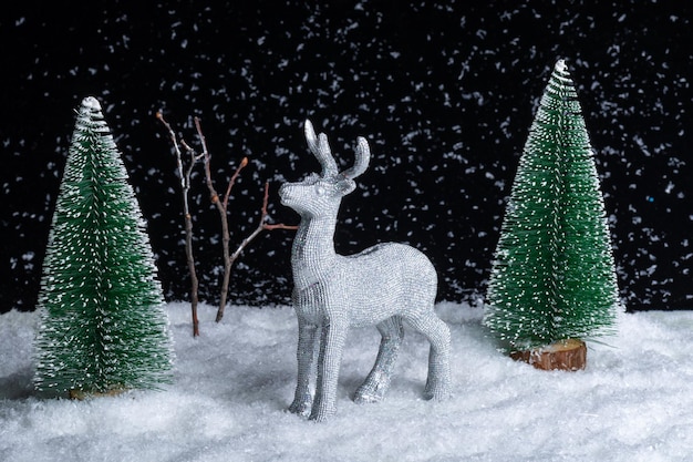 Um cervo prateado brilhante de brinquedo parado na neve na floresta entre as árvores de Natal à noite em um fundo preto