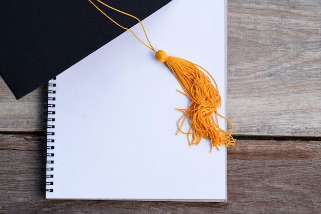 Um chapéu de formatura preto, uma borla de ouro amarelo e um notebook colocado em um conceito de graduação de piso de madeira
