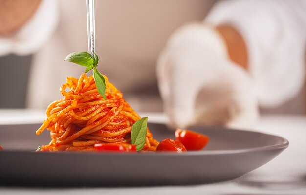 Um chef de luvas e uniforme branco está usando uma pinça para decorar um prato gourmet de macarrão com molho de tomate com tomate cereja, banhando os alimentos