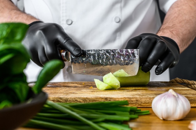 Um chef de luvas pretas está cortando um pepino verde fresco em uma tábua de madeira.
