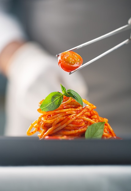 Um chef de uniforme branco e luvas serve tomate cereja no macarrão com molho de tomate com folhas de manjericão com pinça. Culinária gourmet. Alimentos em placas