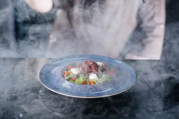 Um chef profissional serve uma salada de tomate e vitela preparada na hora sob um capô de vidro com fumaça espessa