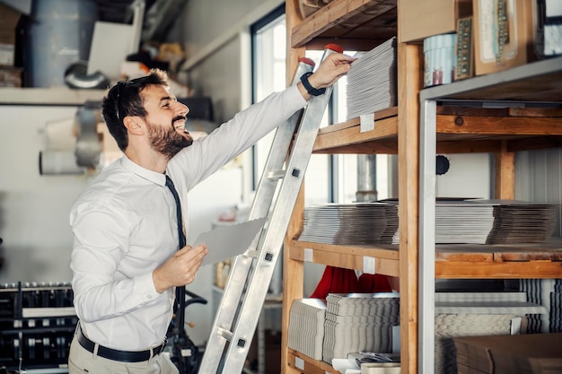 Um chefe feliz em pé nas escadas da gráfica e tirando material das prateleiras