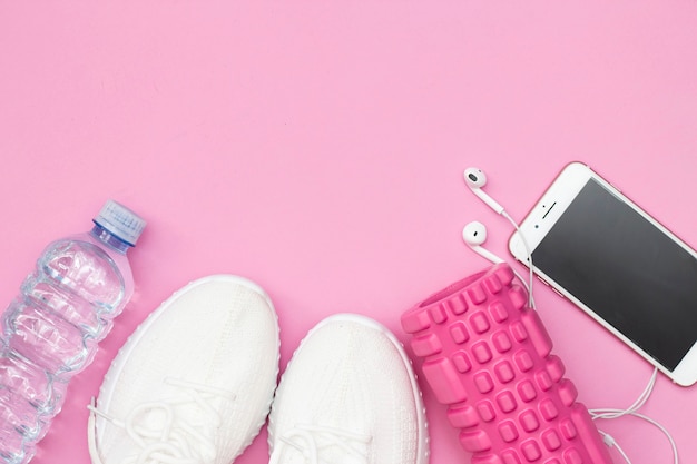 Um conceito de fitness com uma garrafa de água, tênis, um telefone com fones de ouvido, um elástico de fitness roxo, um rolo rosa sobre um fundo rosa. Copie o espaço.