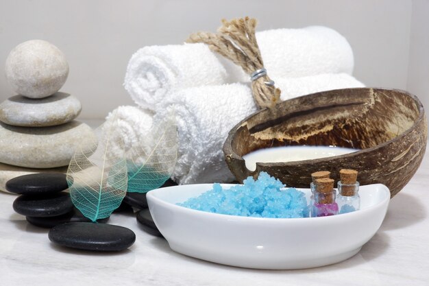 Um conjunto de tratamentos de spa com leite de coco, pedras quentes e sal de banho azul está localizado em uma bancada de mármore branco.