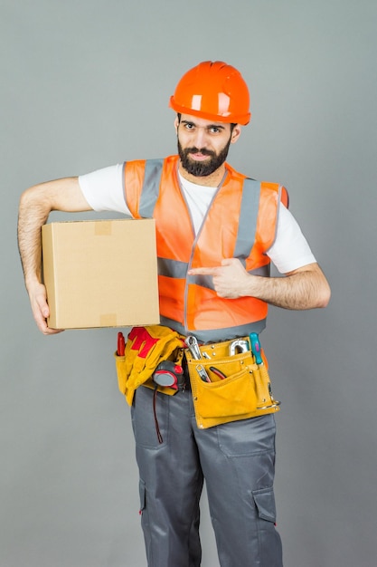 Foto um construtor com um capacete laranja com uma caixa de papelão nas mãos sobre um fundo cinza