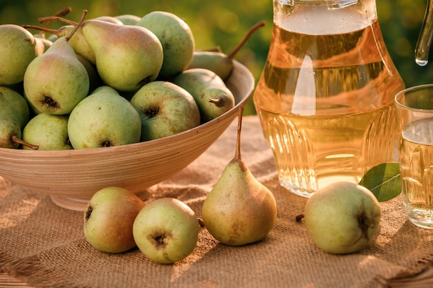 Um copo com suco de pêra e cesta com peras na mesa de madeira com fundo de pomar natural na luz do sol. Composição de frutas vegetarianas