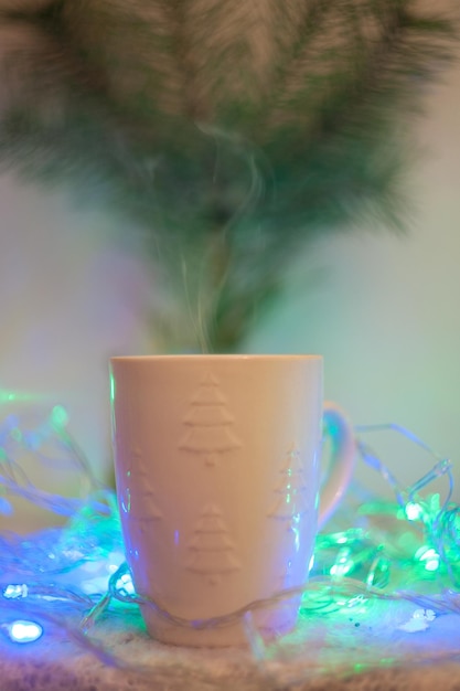 Foto um copo com uma bebida quente contra o fundo de um ramo de árvore de natal e guirlandas desfocadas atmosfera caseira do ano novo