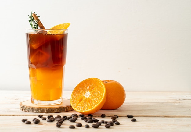 Um copo de café preto americano gelado e uma camada de suco de laranja e limão decorado com alecrim e canela na mesa de madeira