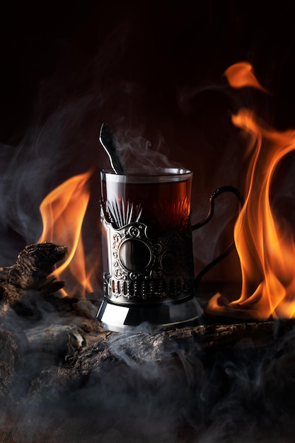 Um copo de chá no porta-copos e jogando línguas e reflexos de chama com fumaça no fundo em troncos de madeira