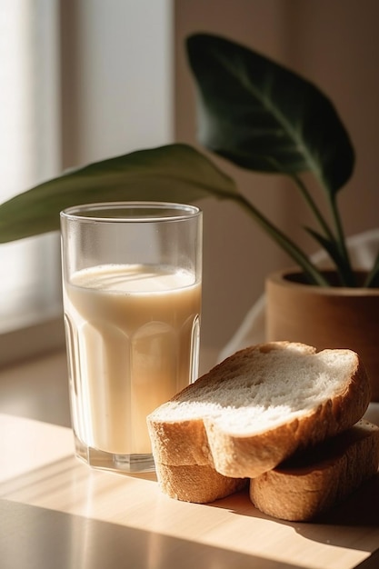 Um copo de leite e um pão sobre uma mesa com uma planta ao fundo.