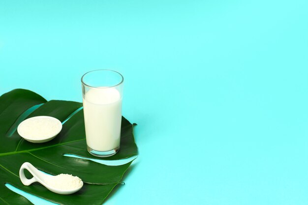 Um copo de leite e uma colher com pó de colágeno ou proteína sobre fundo azul.