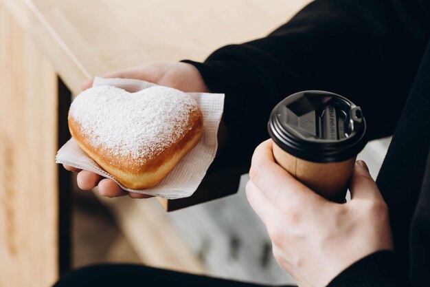 Um copo de papel de café e um donut em forma de coração em mãos masculinas em close-up