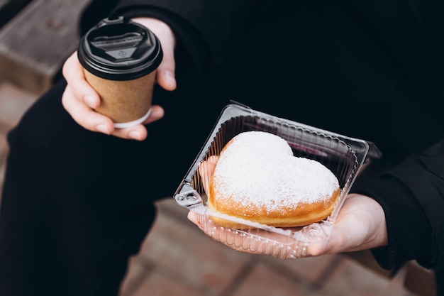 Um copo de papel de café e um donut em forma de coração em mãos masculinas em close-up