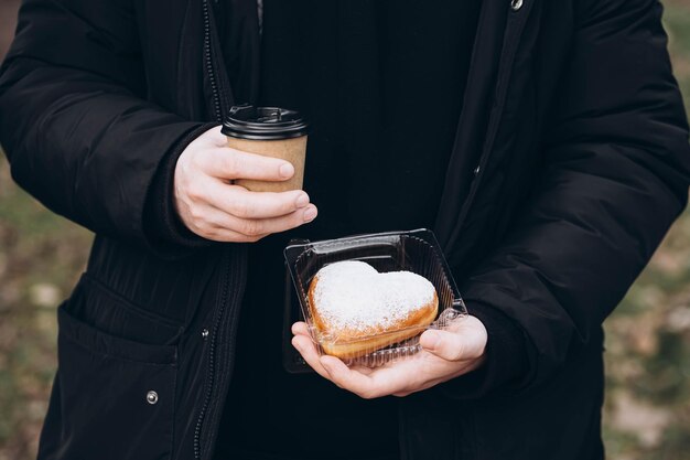Um copo de papel de café e um donut em forma de coração em mãos masculinas em close-up