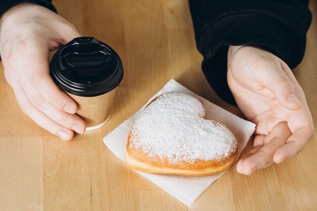 Um copo de papel de café e um donut em forma de coração em mãos masculinas em close-up