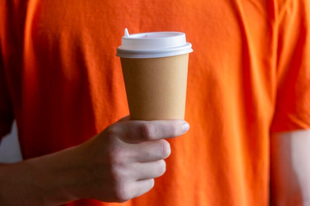 Um copo de papel de café para viagem na mão de um jovem em uma camiseta laranja brilhante.