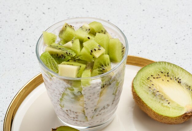Um copo de pudim de sementes de chia coberto com kiwi em cubos fica em um prato com kiwis cortados ao meio e uma colher