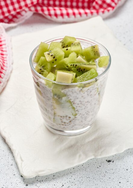 Um copo de pudim de sementes de chia misturado com kiwi em cubos está em uma superfície branca junto com kiwis cortados ao meio