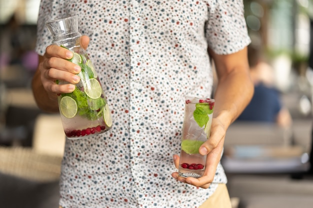Um copo de vidro e uma decantadora com um suco de frutas de verão refrescante de hortelã e limonada de limão cranberry nas mãos de um jovem em uma camisa de manga curta.
