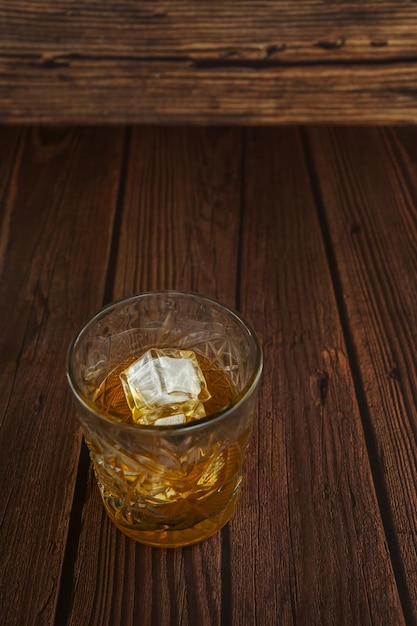 Um copo de vidro transparente cheio de licor de mel com alguns cubos de gelo ao lado de uma garrafa sobre uma mesa de madeira