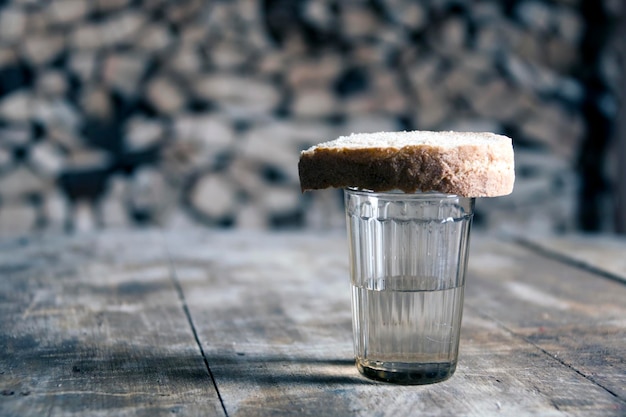 Um copo de vodka e um pedaço de pão em uma mesa de madeira e lenha atrás