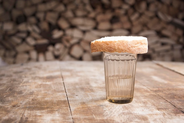 Um copo de vodka e um pedaço de pão em uma mesa de madeira e lenha atrás