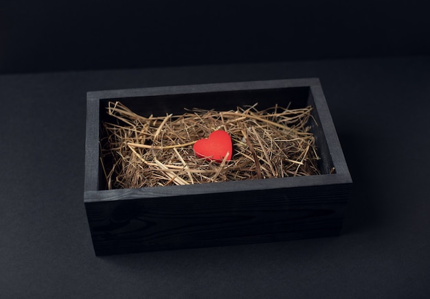 Um coração vermelho dentro de uma caixa de madeira preta com feno (grama seca) na superfície escura