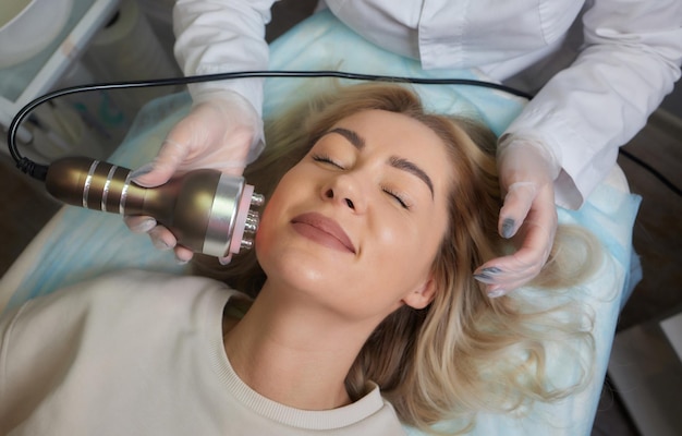 Um cosmetologista em luvas de borracha faz o procedimento de rflifting no rosto No fundo, o dispositivo de cosmetologia Rf Close up Vista lateral O conceito de cosmetologia profissional e rejuvenescimento